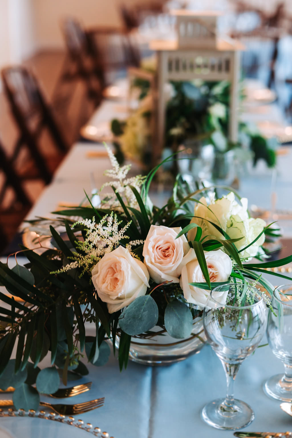 Elegant Coastal Glam Wedding Reception Table Decor with Low Blush Rose and Natural Greenery Centerpiece in Silver Bowl, with Blue Linens and Antique Old Florida Hurricane Lanterns