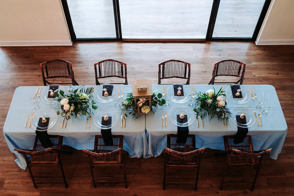 Coastal Glam Intimate Wedding Reception Decor with Long Feasting Table with Blue Linens, Low Natural Greenery and Hurricane Lantern Centerpiece, Bamboo Folding Chairs, and Gold Chargers and Flatware | Tampa Bay Waterfront Wedding Venue Beso Del Sol Resort | Coast to Coast Event Rentals