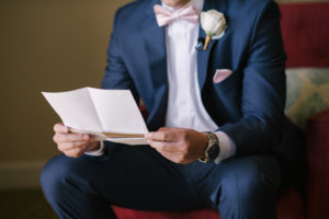 Groom Reading Letter from the Bride Wedding Portrait wearing Navy Suit, Blush Bowtie, and White Rose Boutonnière