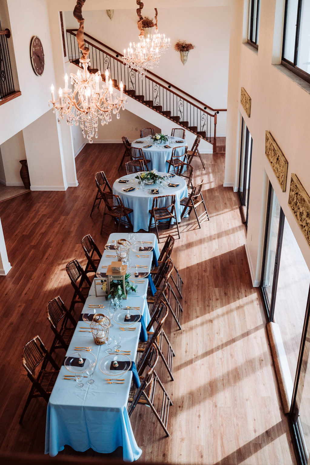 Coastal Glam Wedding Reception Long Feasting Table with Blue Linens, Low Natural Greenery and Hurricane Lantern Centerpiece, Bamboo Folding Chairs, and Gold Chargers and Flatware | Tampa Bay Waterfront Wedding Venue Beso Del Sol Resort | Wedding Rentals Coast to Coast Event Rentals