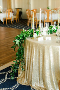 Old Florida Inspired White, Gold, and Green Wedding Reception Table Decor with Glitter Tablecloth, White Rose and Greenery Garland Centerpiece, and Elegant Candlestick holders | Clearwater Beach Wedding Reception Venue Carlouel Yacht Club | Tampa Bay Wedding Planner Glitz Events