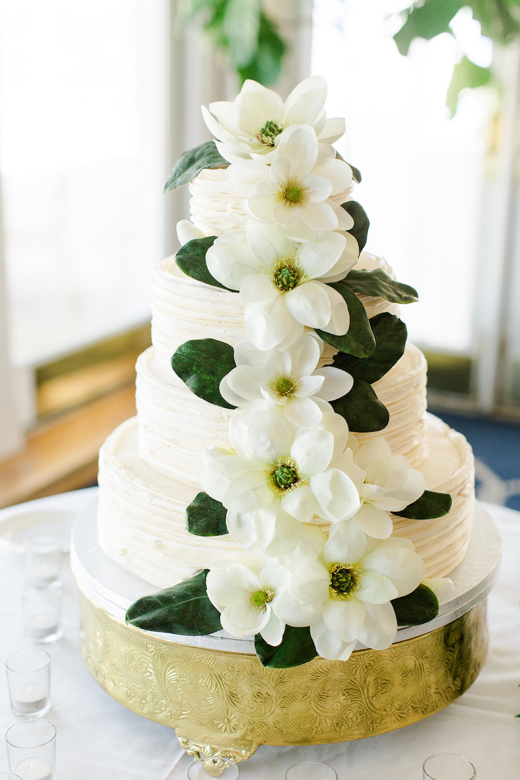 Four Tiered Round White Wedding Cake with White Magnolia and Greenery Flowers on Gold Cake Tray