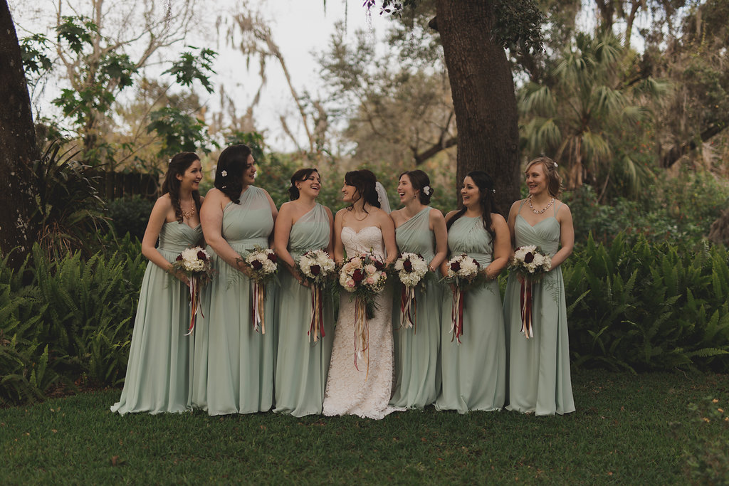 Outdoor Garden Bridal Party Portrait, Bride in Strapless A Line Floral Lace Stella York Wedding Dress, Bridesmaids in Mismatched Sage Green Floor Length Dresses, with Ivory, Blush, and Dark Red Bouquets with Greenery and Long Champagne and Maroon Ribbons | Tampa Bay Wedding Photographer Stacy Paul Photography | Tampa Bay Rustic Wedding Venue Cross Creek Ranch