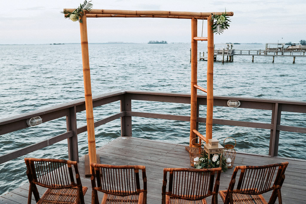 Intimate Coastal Themed Waterfront Wedding Ceremony Decor with Bamboo Arch and Folding Chairs, Vintage Hurricane Lanterns with Natural Tropical Greenery and White Florals | Tampa Bay Wedding Venue Beso Del Sol Resort