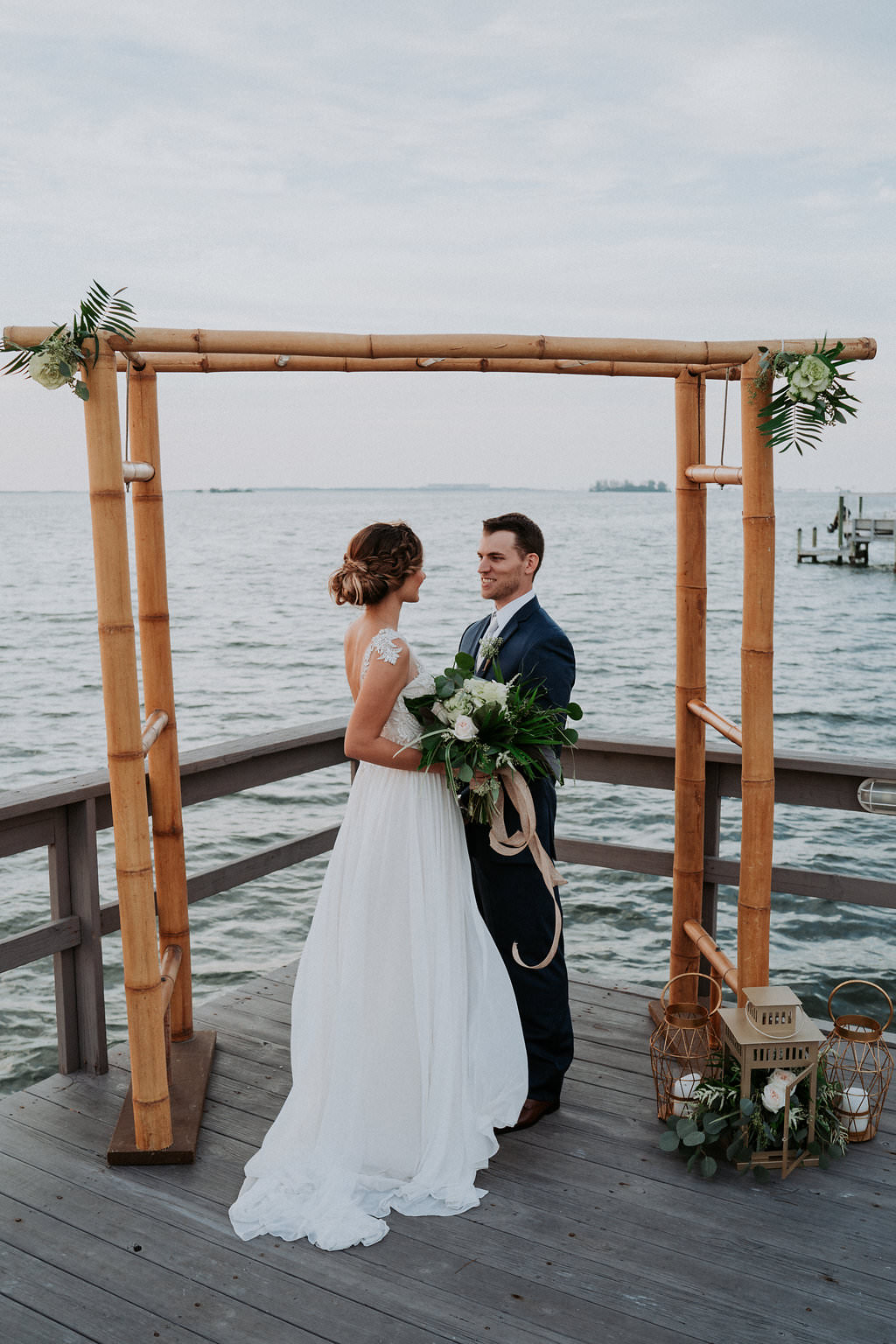 Coastal Themed Waterfront Wedding Ceremony Portrait with Bamboo Arch with Vintage Hurricane Lanterns and Natural Tropical Greenery and White Florals | Dunedin Waterfront Hotel Wedding Venue Beso Del Sol Resort | Tampa Bay Wedding Photographer Grind and Press Photography