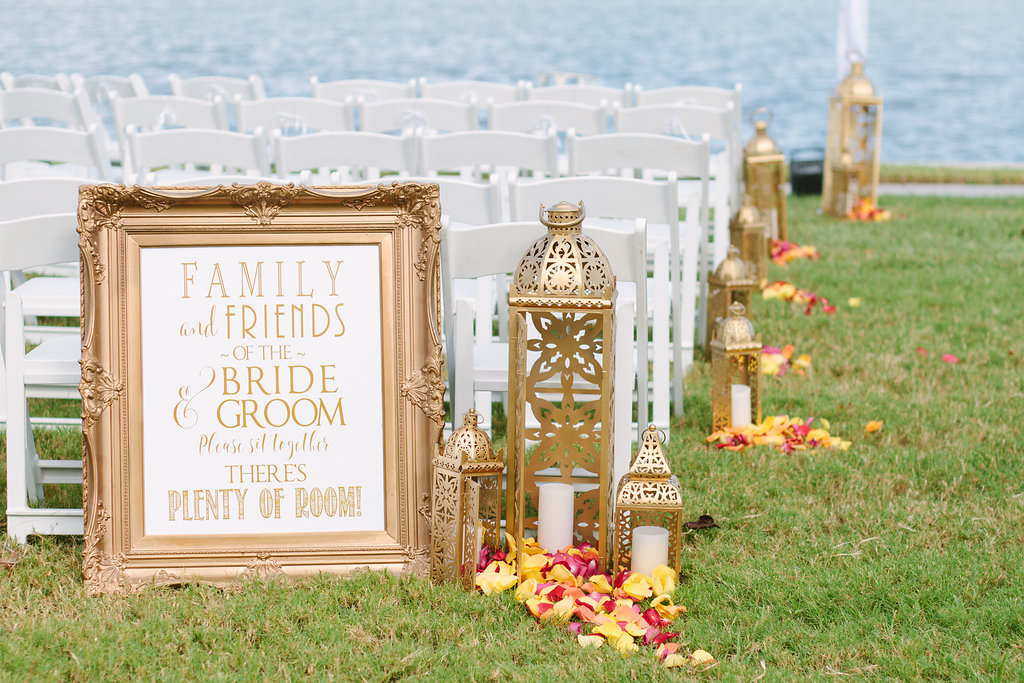 Outdoor Waterfront Whimsical Wedding Ceremony Decor with Ornate Gold Frame Welcome Sign and Gold Moroccan Lanterns of Varying Heights with Yellow and Red Petals and White Folding Chairs | Tampa Bay Wedding Planner Special Moments Event Planning