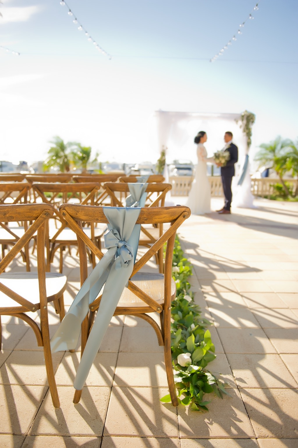 Outdoor Waterfront French Countryside Inspired Wedding Ceremony with Wooden Cross Back Chairs with Wide Light Blue Ribbon, Greenery Garland Aisle, and White Draped Ceremony Arch | Tampa Bay Wedding Planner Kelly Kennedy Weddings and Events | Tampa Waterfront Outdoor Wedding Venue Westshore Yacht Club