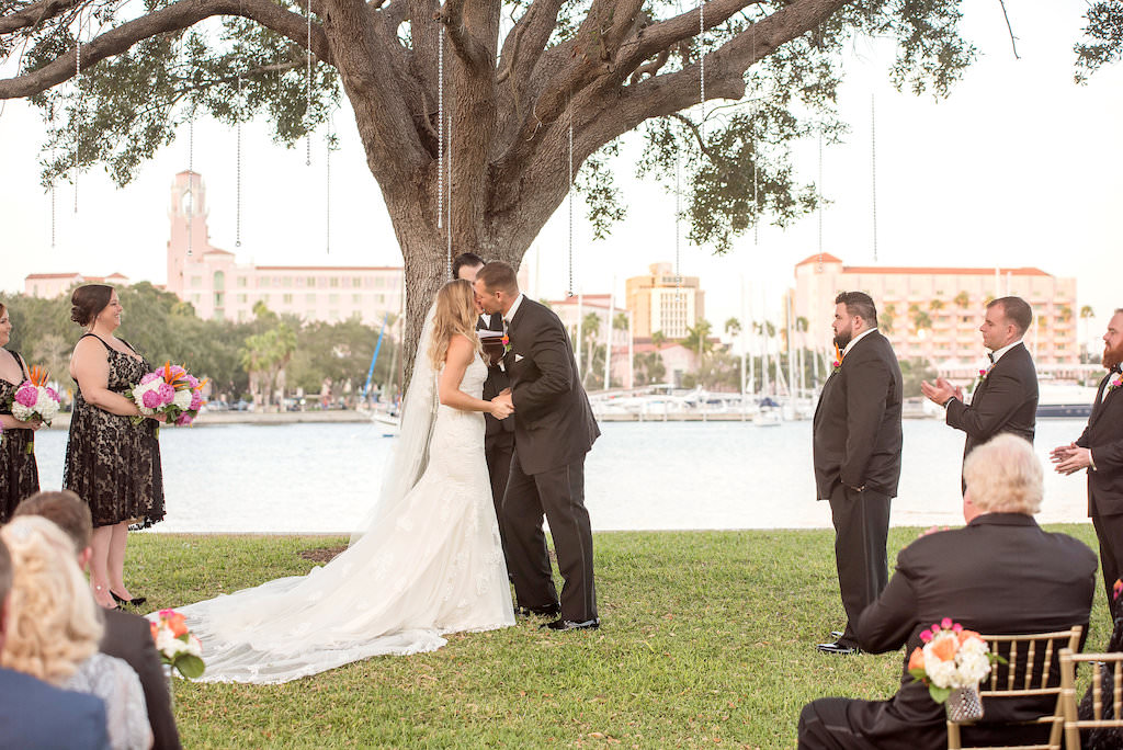 Outdoor Waterfront Wedding Ceremony Portrait, Groom in Black Tuxedo with Orange and Pink Tropical Bird of Paradise Boutonniere, Hanging Chrystal Beads Decor from Tree, and Gold CHiavari Chairs | Bridesmaids in Black Lace Knee Length Dresses with Pink, Orange and White Bouquets | Downtown St Pete Historic Outdoor Wedding Ceremony Venue The St Petersburg Museum of History | Tampa Bay Wedding Photographer Kristen Marie Photography