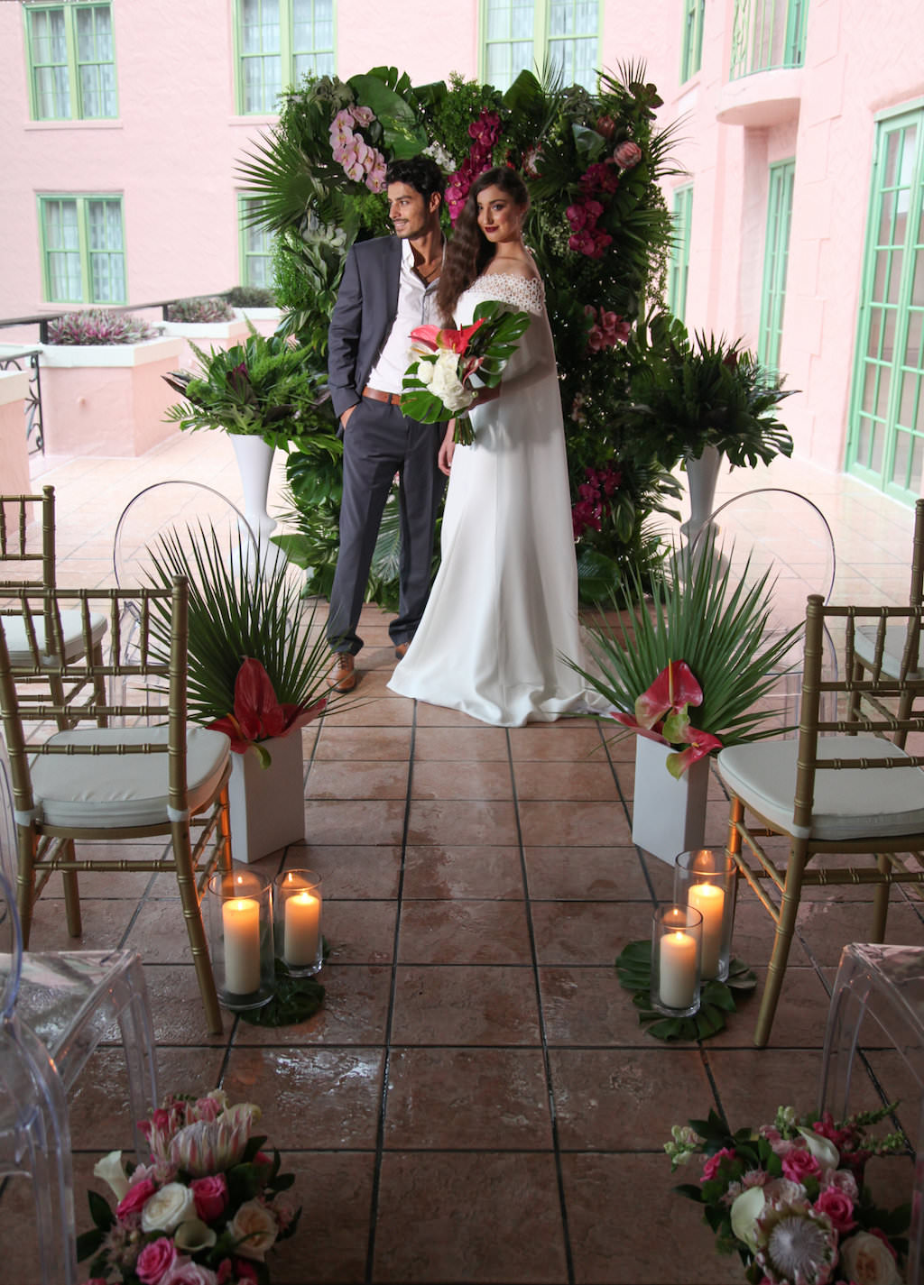 Intimate Hotel Courtyard Tropical Latin Cuban Inspired Wedding Ceremony, with Gold Chiavari Chairs, Tall Pillar Candles in Glass Hurricane Lanterns, Magenta, Pink and White Tropical Florals with Fern and Palm Frond Greenery, With Clear Acrylic Oval Backed Chairs | Tampa Bay Wedding Rentals A Chair Affair | Downtown St Pete Venue Vinoy Renaissance