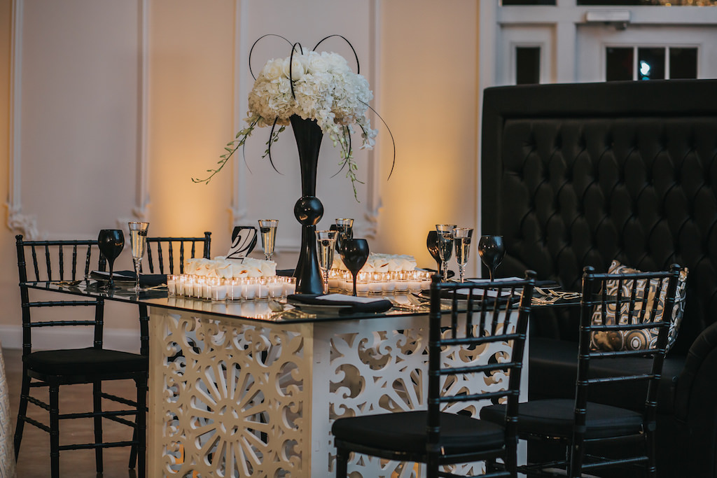Modern, Sophisticated Black and White Wedding Reception Decor with Tall White Hydrangea Centerpiece in Geometric Black Vase, Black Wine Glasses, Stylish White Carved Wooden Table with Glass Tabletop, and Black Chiavari Chairs | Tampa Bay Wedding Rentals A Chair Affair | Wedding Planning UNIQUE Weddings and Events