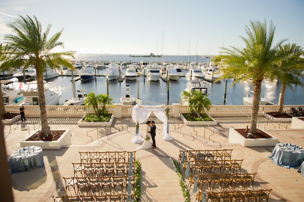 Outdoor Waterfront French Countryside Inspired Wedding Ceremony with Wooden Cross Back Chairs with Wide Light Blue Ribbon, Greenery Garland Aisle, and White Draped Ceremony Arch | Tampa Bay Wedding Planner Kelly Kennedy Weddings and Events | Tampa Waterfront Outdoor Wedding Venue Westshore Yacht Club