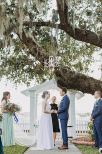 Outdoor Garden Wedding Ceremony with Folding White Chairs with Greenery and Sage Green Ribbon Florals and Wooden Unplugged Ceremony Welcome Sign with White Painted Script | Bradenton Wedding Venue Palmetto Riverside Bed and Breakfast | Sarasota Wedding Planner Jennifer Matteo Event Planning