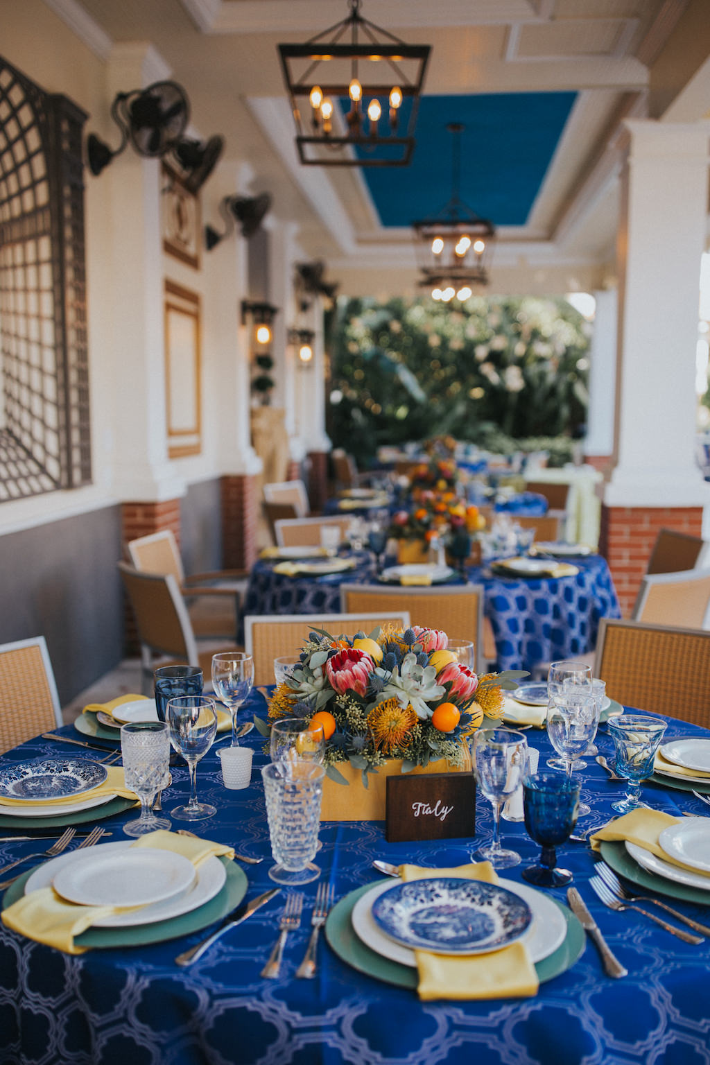 Mediterranean Inspired Wedding Reception Round Table Decor with Low Yellow, Orange, Pink and Succulent Greenery Centerpiece, Wooden Table Number European Country Names Sign with White Script, Sage Green Chargers and Yellow Napkins, Cobalt Blue Patterned Tablecloth, and Blue Painted China Plates | Sarasota Wedding Planner Jennifer Matteo Event Planning | Over the Top Rental Linens