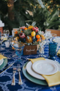 Mediterranean Inspired Wedding Reception Round Table Decor with Low Yellow, Orange, Red and Succulent Greenery Centerpiece with Lemons and Persimmon, Wooden Table Number European Country Names Sign with White Script, Sage Green Chargers and Yellow Napkins, Cobalt Blue Patterned Tablecloth, and Blue Painted China Plates | Sarasota Wedding Planner Jennifer Matteo Event Planning | Over the Top Rental Linens