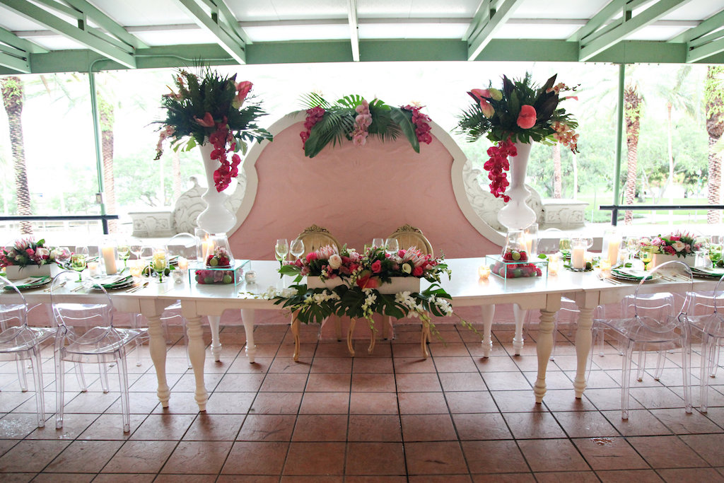 Outdoor Hotel Courtyard Old World Havana Nights Cuban Inspired Wedding Reception with Long White Vintage Feasting Table with Clear Acrylic Oval Backed Chairs, Extra Tall Pink Orchid and Fern Greenery with Palm Fronds Centerpiece in White Vase, Low Centerpiece with Monsterra Palm Leaves and Florals in White Boxes, and Pillar Candles | Tampa Bay Wedding Rentals A Chair Affair | St Pete Venue Vinoy Renaissance