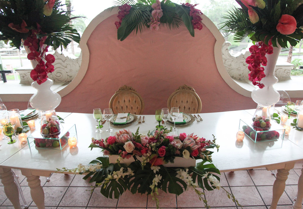 Outdoor Hotel Courtyard Old World Havana Nights Cuban Inspired Wedding Reception with Long White Vintage Feasting Table with Clear Acrylic Oval Backed Chairs, Extra Tall Pink Orchid and Fern Greenery with Palm Fronds Centerpiece in White Vase, Low Centerpiece with Monsterra Palm Leaves and Florals in White Boxes, and Pillar Candles | Tampa Bay Wedding Rentals A Chair Affair | St Pete Venue Vinoy Renaissance