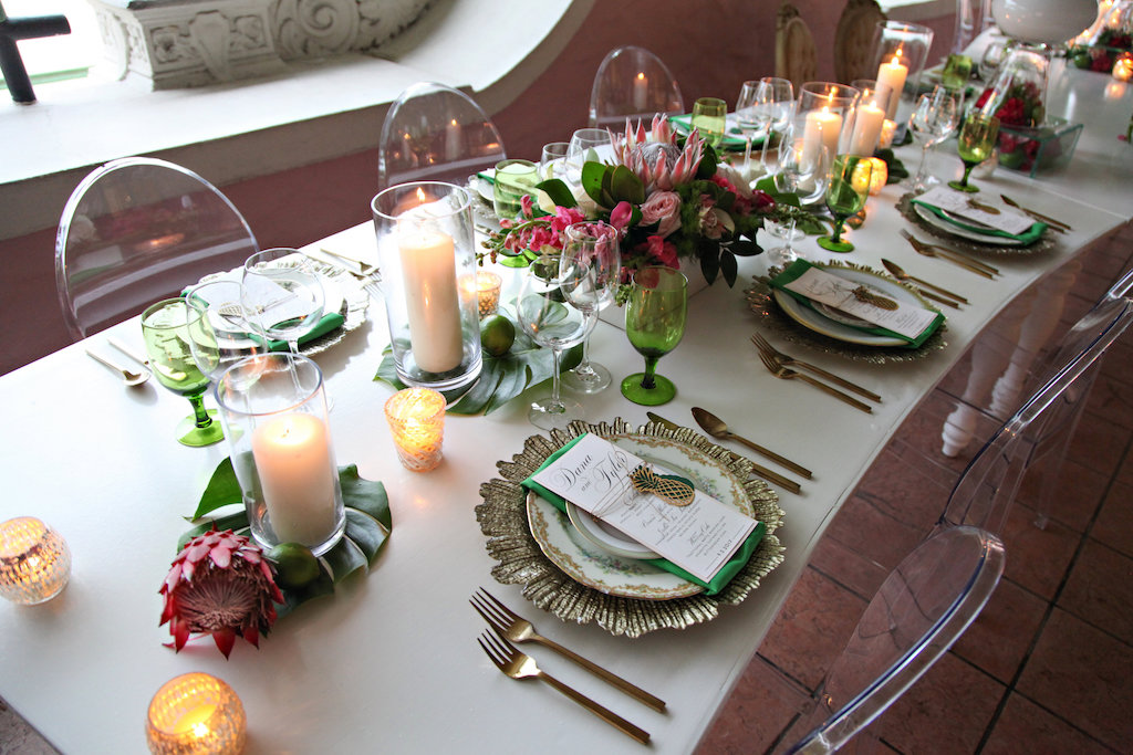 Old World Havana Inspired Wedding Reception Long White Curved Feasting Table with Pillar Candles on Monsterra Palm Fronds with Protea, Green Satin LInens, Pink LIly and White Orchid Low Centerpiece in White Box, Gold Chargers with Vintage China Place Settings and Brushed Flatware, and Oval Backed Clear Acrylic Chairs | St Pete Wedding Rentals A Chair Affair