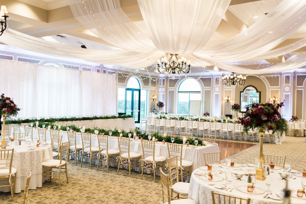 Whimsical Ballroom Wedding Reception with Gold Chiavari Chairs and Long White Feasting Tables with Greenery Garland Table Runner Centerpiece, Tall Magenta and Purple with Greenery Centerpiece in Tall Gold Vase, and White Ceiling Draping | Sarasota Country Club Wedding Venue Lakewood Ranch Golf & Country Club