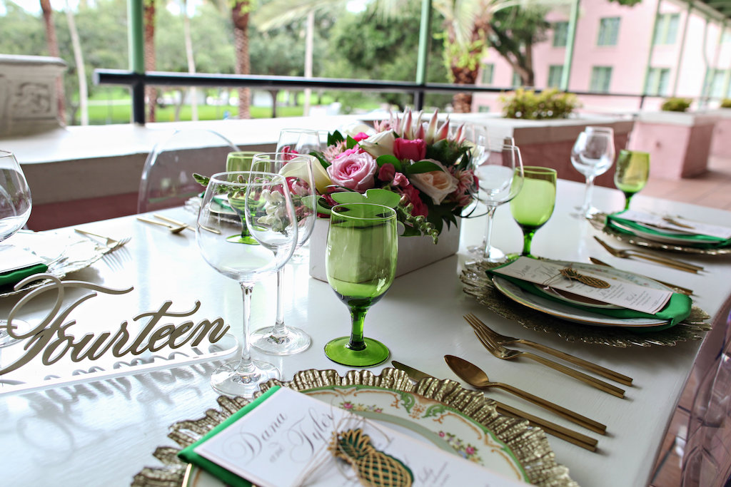 Old World Havana Inspired Wedding Reception Long White Feasting Table with Pink Lily and White Orchid and Protea Low Centerpiece in White Box, Gold Chargers with Vintage China Place Settings and Brushed Flatware, Green Linens and Colored Drinking Glasses, Pineapple Favor, Stylish Gold Table Number, and Oval Backed Clear Acrylic Chairs | St Pete Wedding Rentals A Chair Affair