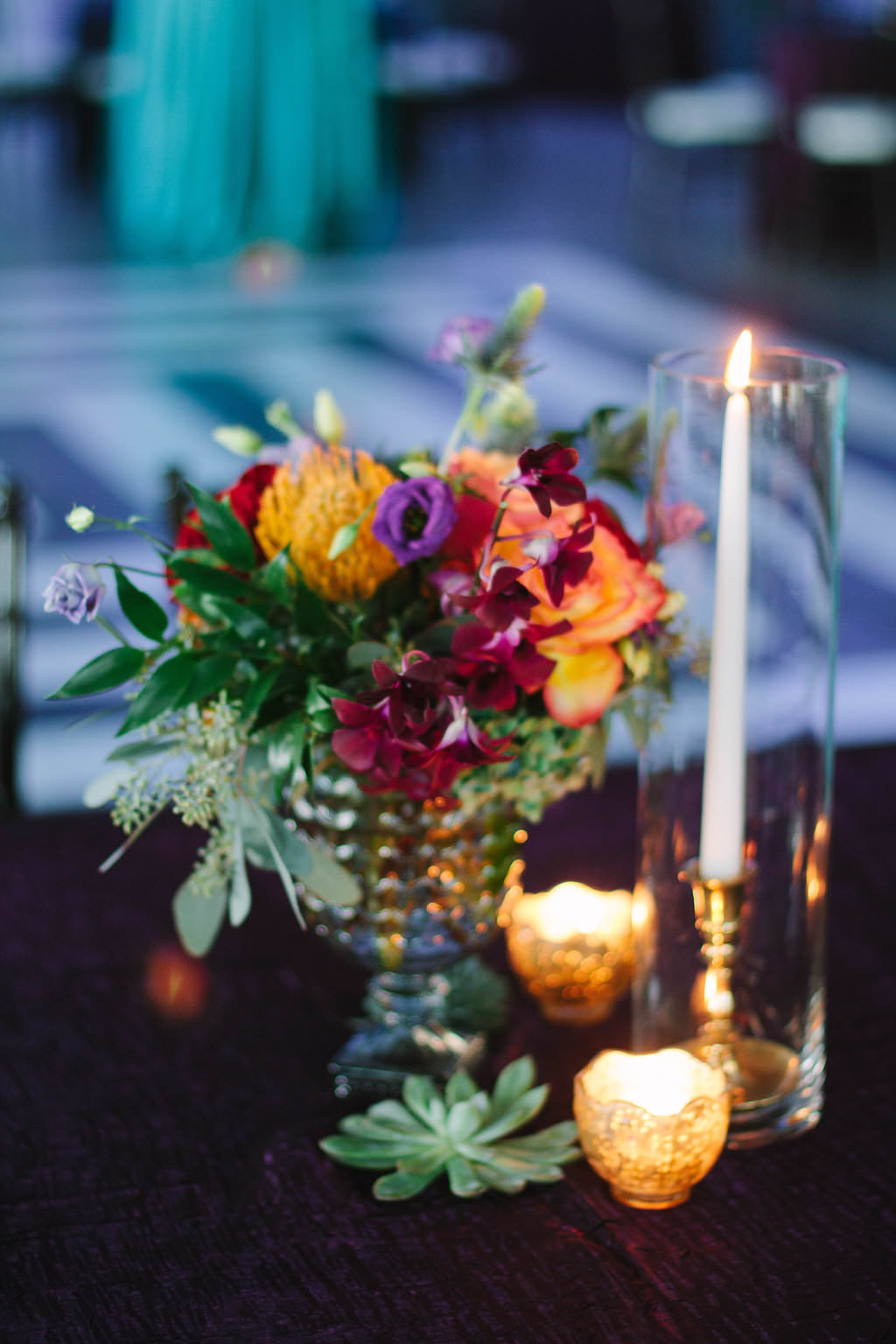Whimsical Jewel Tone Wedding Reception Table Decor with Small Dark Red, Yellow, and Purple Flowers with Greenery and Succulent Centerpiece and Gold Tea Lights and Taper Candle in Dainty Gold Candleholder in Glass Vase