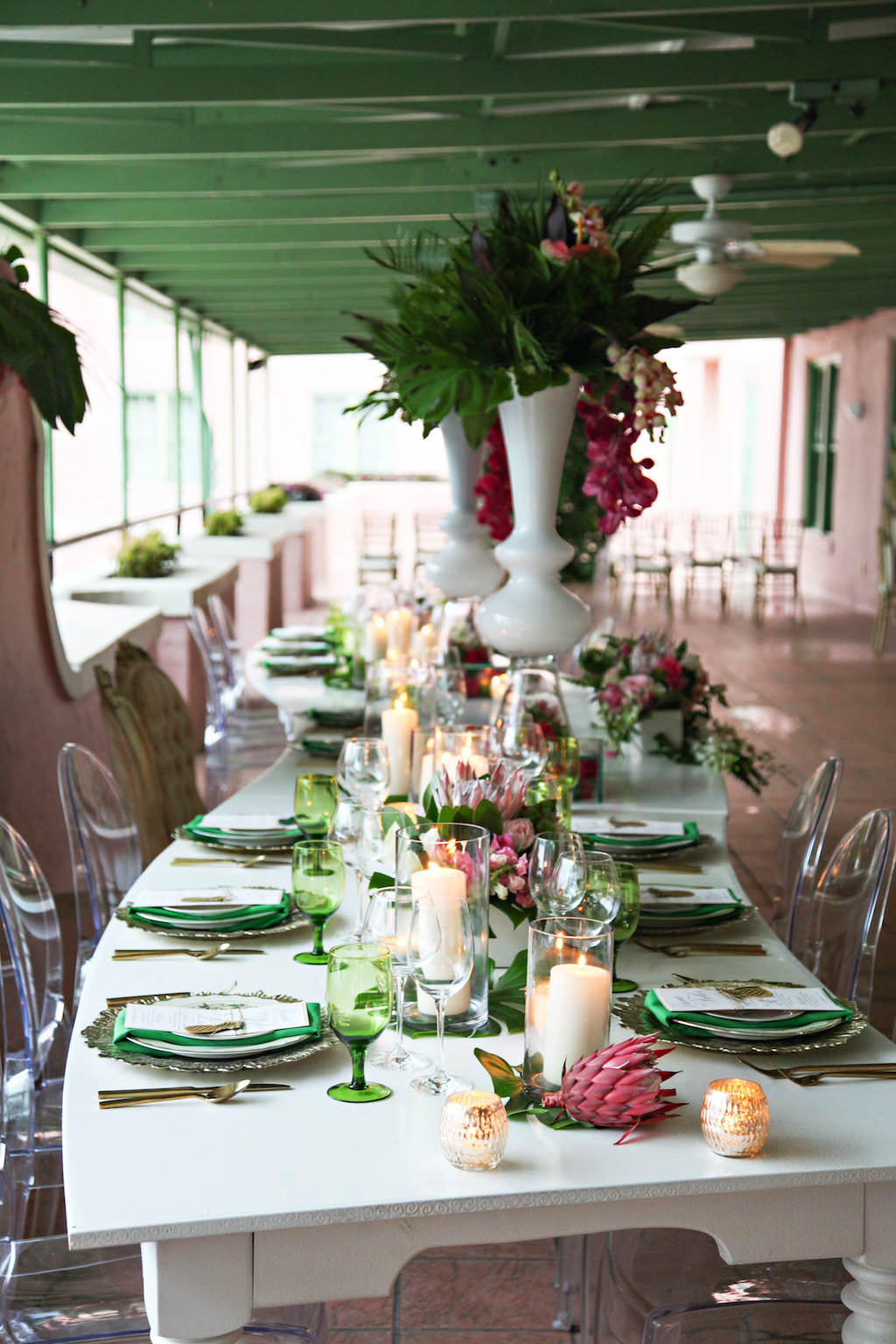 Outdoor Hotel Courtyard Old World Havana Inspired Wedding Reception with Long White Vintage Feasting Table with Clear Acrylic Oval Backed Chairs, Extra Tall Pink Orchid and Fern Greenery with Palm Fronds Centerpiece in White Vase, Low Centerpiece with Monsterra Palm Leaves and Florals in White Boxes, Colored Drinking Glasses and Green Satin Napkins, and Pillar Candles in Hurricane Votives | Tampa Bay Wedding Rentals A Chair Affair | St Pete Venue Vinoy Renaissance