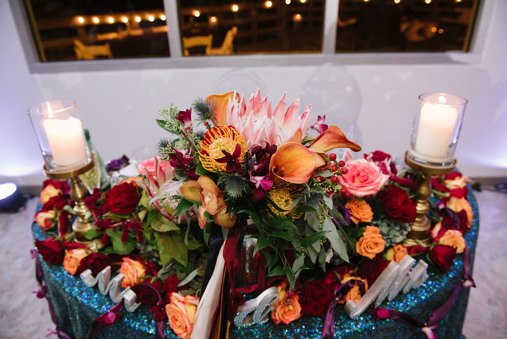 Whimsical Jewel Tone Wedding Reception Sweetheart Table with Large Orange, Dark Red, Purple and Pink Centerpiece with Ribbon and Greenery and Succulents and Thick Gold Candleholders with Pillar Candles and Glittering Emerald Green Tablecloth | Tampa Bay Wedding Planner Special Moments Event Planning