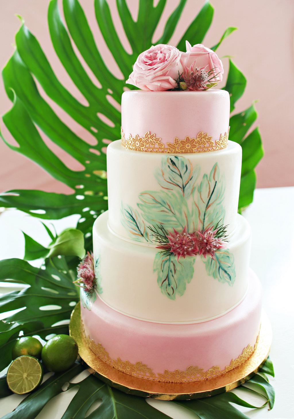 Four Tiered Tropical Havana Inspired Wedding Cake with PInk Icing, Painted Leaf Details and Pink Roses and Gold Floral Details, on Gold Cakestand with Monsterra Palm Frond and Limes