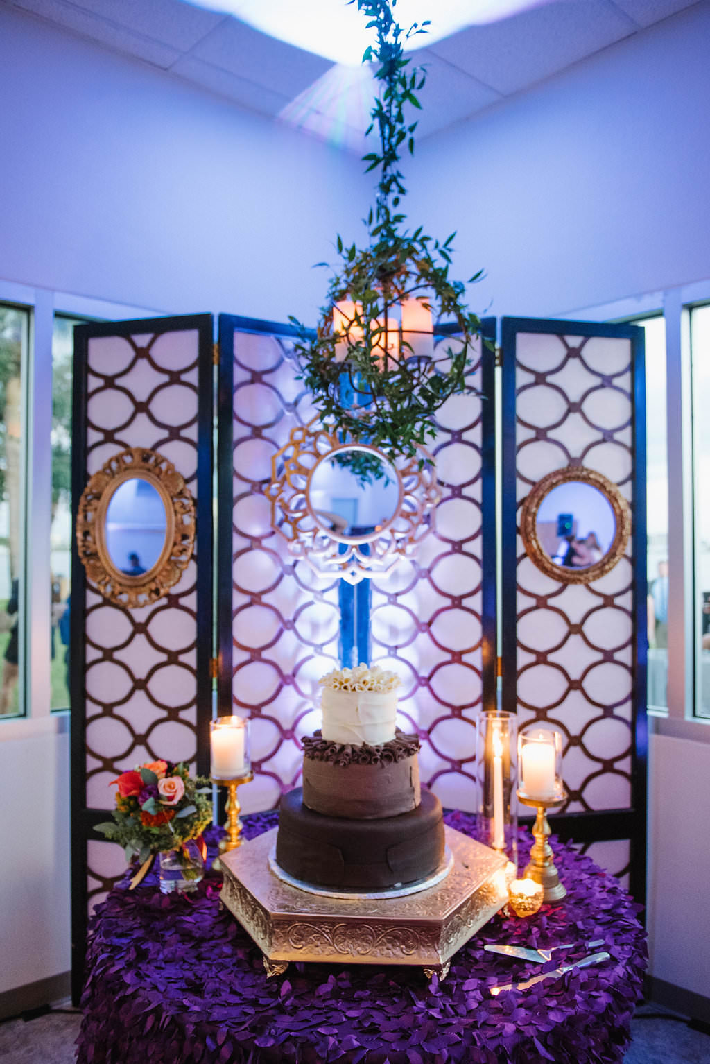 Whimsical Jewel Tone Wedding Dessert Table with Three Tier Round White, Milk, and Dark Chocolate Wedding Cake on Gold Octagonal Cake Stand on Purple Textured Linen with Pillar Candles, Hanging Greenery Wreath, and Vintage Pink, Gold and Black Screen | Tampa Bay Wedding Dessert Bakery A Piece of Cake | Clearwater, FL Wedding Planner Special Moments Event Planning