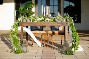 French Countryside Outdoor Wedding Reception Sweetheart Table with Vintage Wooden Chairs, Long White Runner with Greenery Garland Centerpiece, and Blue Colored Drinking Glasses | Tampa Wedding Planner Kelly Kennedy Weddings & Events | Tampa Bay Waterfront Wedding Venue The Westshore Yacht Club