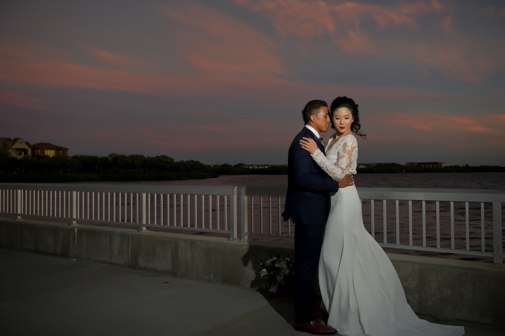 Waterfront Sunset Bride and Groom Wedding Portrait, Groom with Red and White Plaid Bowtie and Blue Suit, Bride in V Neck Lace Long Sleeve Daalarna Couture Wedding Dress | Tampa Wedding Photographer Andi Diamond Photography | Tampa Bridal Wedding Dress Shop The Bride Tampa