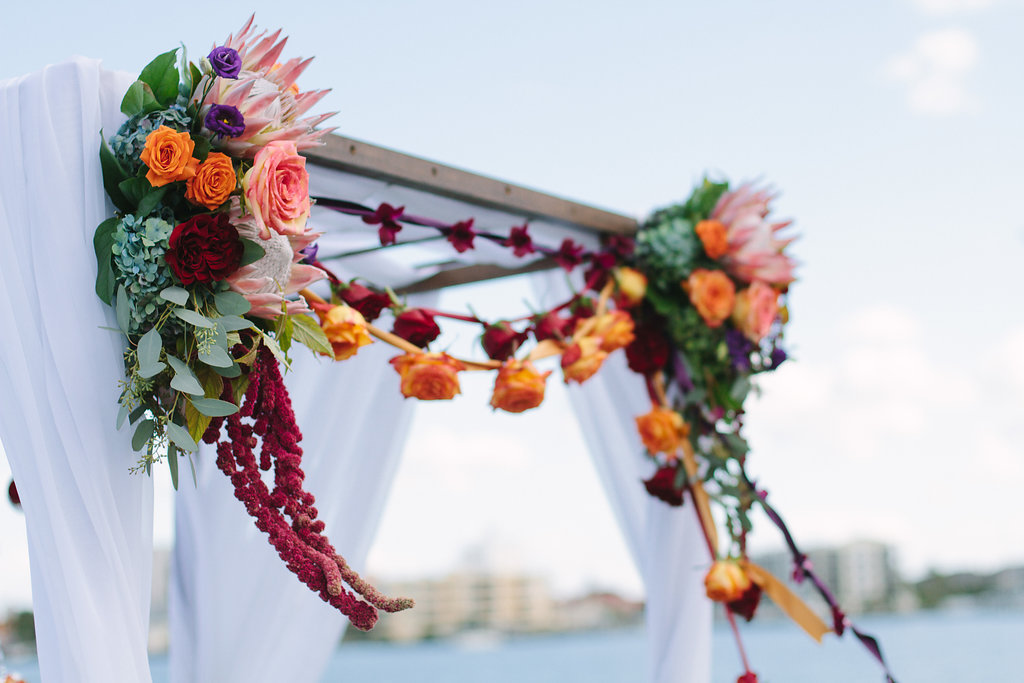 Outdoor Whimsical Jewel Tone Beach Wedding Ceremony Arch with Magenta, Dark Red, Orange, Pink, and Purple Roses with Greenery And Ribbon Garlands | Tampa Bay Wedding Planner Special Moments Event Planning