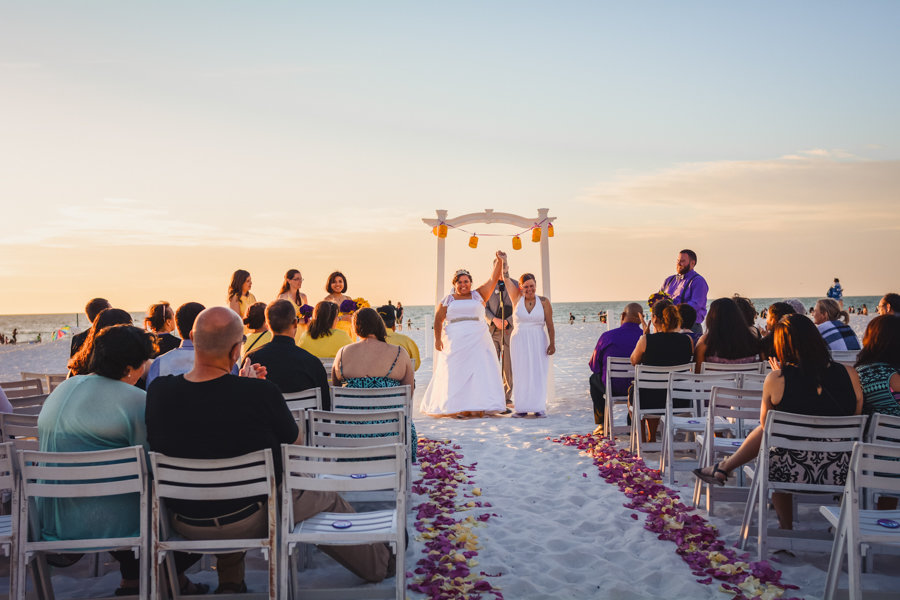 Same-Sex Gay Beach Wedding Ceremony Portrait, with White Wooden Arch with Yellow Paper Lanterns, White Folding Chairs, and Purple and Cream Flower Petal Aisle, Bridesmaids in Yellow with Purple Bouquets | Beach Hotel Wedding Venue Hilton Clearwater Beach