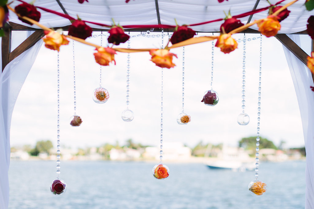 Outdoor Whimsical Jewel Tone Beach Wedding Ceremony Arch with Orange and Dark Red Rose and Ribbon Garlands, White Draping, and Hanging Glass Bead Strands with Glass Ball Hanging Vases | Tampa Bay Wedding Planner Special Moments Event Planning