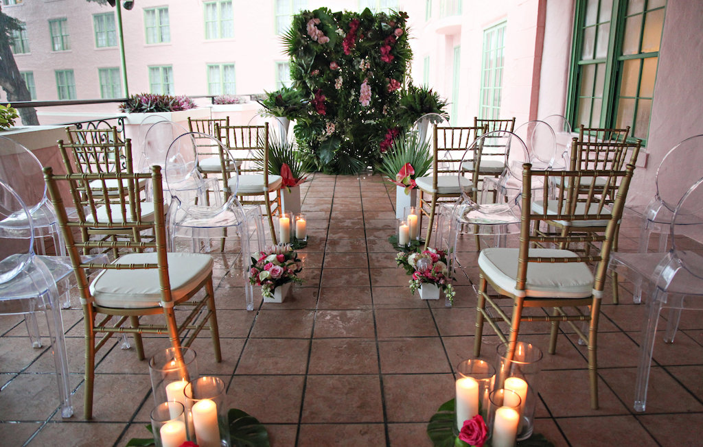 Intimate Hotel Courtyard Tropical Latin Cuban Inspired Wedding Ceremony, with Gold Chiavari Chairs, Tall Pillar Candles in Glass Hurricane Lanterns, Magenta, Pink and White Tropical Florals with Fern and Palm Frond Greenery, With Clear Acrylic Oval Backed Chairs | Tampa Bay Wedding Rentals A Chair Affair | Downtown St Pete Venue Vinoy Renaissance