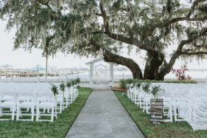 Outdoor Garden Wedding Ceremony with Folding White Chairs with Greenery and Sage Green Ribbon Florals and Wooden Unplugged Ceremony Welcome Sign with White Painted Script | Bradenton Wedding Venue Palmetto Riverside Bed and Breakfast | Sarasota Wedding Planner Jennifer Matteo Event Planning