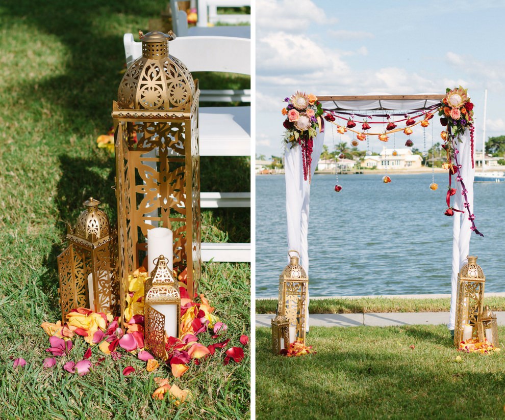 Outdoor Waterfront Whimsical Jewel Tone Wedding Ceremony Decor with Tall Moroccan Lanterns and Yellow and Red Flower Petals, and Ceremony Arch with Red and Gold Rose Garlands with Ribbon and White Draping | Tampa Bay Wedding Planner Special Moments Event Planning