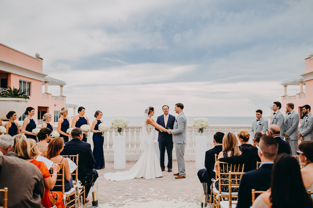 Rooftop Wedding Ceremony Portrait, Bridesmaids in Navy Blue Column Dresses, Groomsmen in Gray Suits with Navy Blue Bowties, with Gold Pillar Candles in Glass Cylinder Hurricane Lanterns, and Gold and White Chiavari Chairs | Tampa Bay Wedding Photographer Rad Red Creative | Waterfront Hotel Wedding Venue Hyatt Regency Clearwater Beach