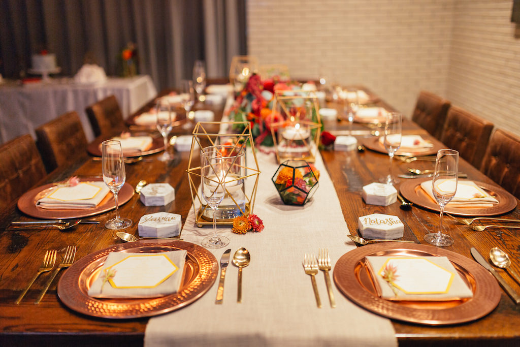 Intimate Wedding Reception Table Decor with Floating Votive Candles in Brass Geometric Holders with Red and Orange Floral Centerpiece with Greenery on Simple Linen Runner with Marble Place Cards and Gold Flatware and Copper Chargers on Wooden Table | Tampa Wedding Planner UNIQUE Weddings and Events | Rentals Over The Top Linens | Venue The Epicurean