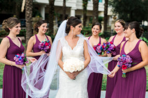 Outdoor Bridal Party Portrait, Bride with White Rose Bouquet in V Neck Lace Maggie Sottero Wedding Dress, Bridesmaids in Davids Bridal Fuchsia V Neck Dresses with Purple, Red, and Blue Bouquets