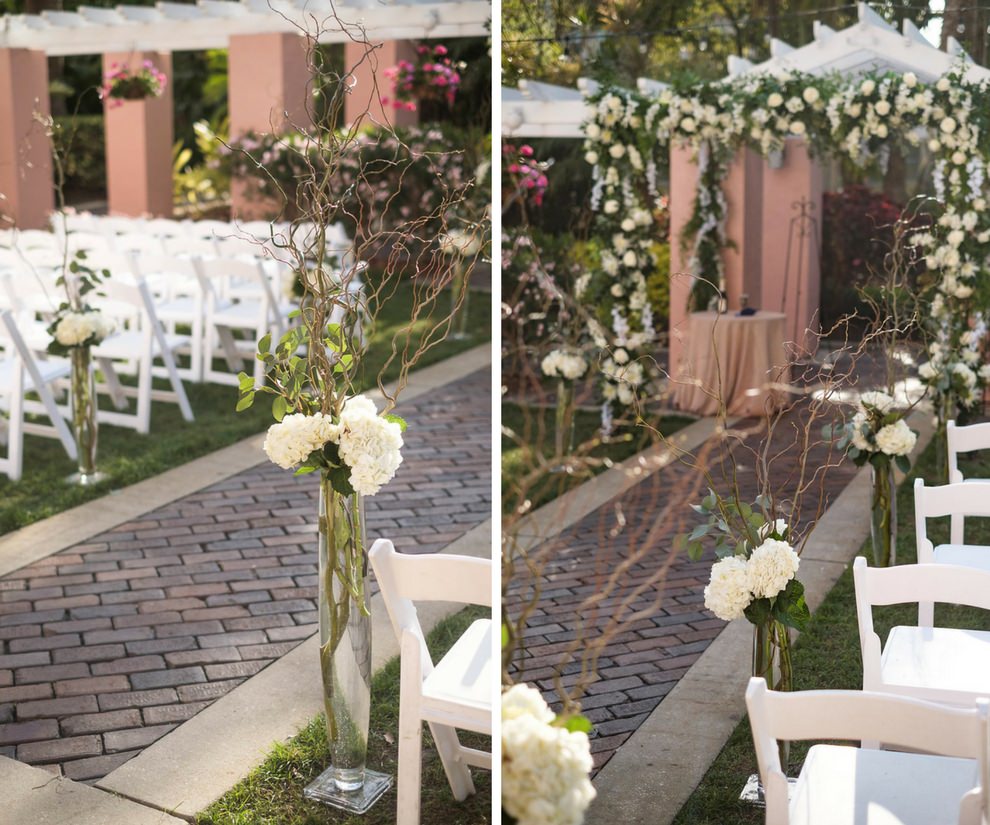 Outdoor Courtyard Jewish Wedding Ceremony Decor with White Folding Chairs, White And Greenery Garland Chuppah Wedding Arch, and Tall Hydrangea and Greenery with Natural Branches Flower Arrangements in Glass Vases | St Pete Historic Hotel Wedding Venue The Vinoy