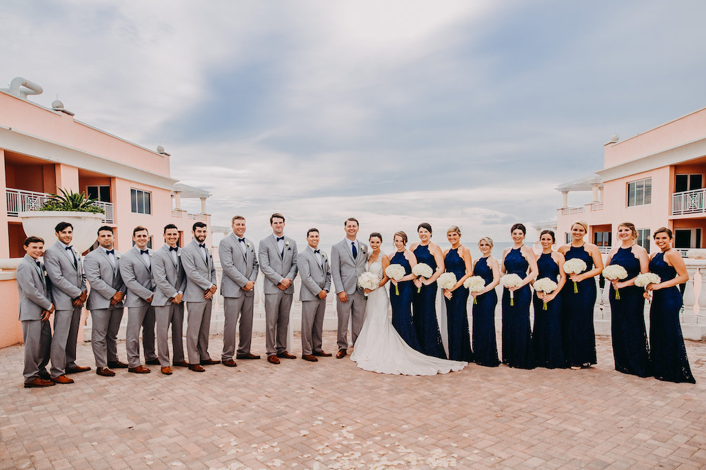 Outdoor Rooftop Wedding Party Portrait, Bridesmaids in Halter Navy Blue Column Lulu's Zenith Dresses, Bride in Lace Cap Sleeve A Line Sincerity Bridal Wedding Dress, with White Floral Bouquet, and Groomsmen in Light Gray Suits with Blue Bowties and Brown Shoes | Tampa Bay Wedding Photography Rad Red Creative | Waterfront Hotel Wedding Venue Hyatt Regency Clearwater Beach