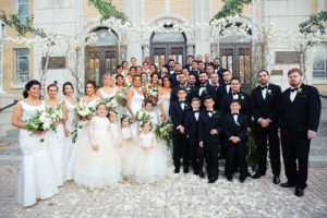 Outdoor Wedding Party Portrait, Bridesmaids in Floorlength White Hayley Paige Bella Bridesmaids Dresses, Groomsen in Black Tuxedos with White and Greenery Boutonniere, Flower Girls in Blush Pink Dresses with Layered Skirts, under hanging White Flowers, Greenery, and Wild Branch Arches | Tarpon Springs Venue St Nicholas Greek Orthodox Church