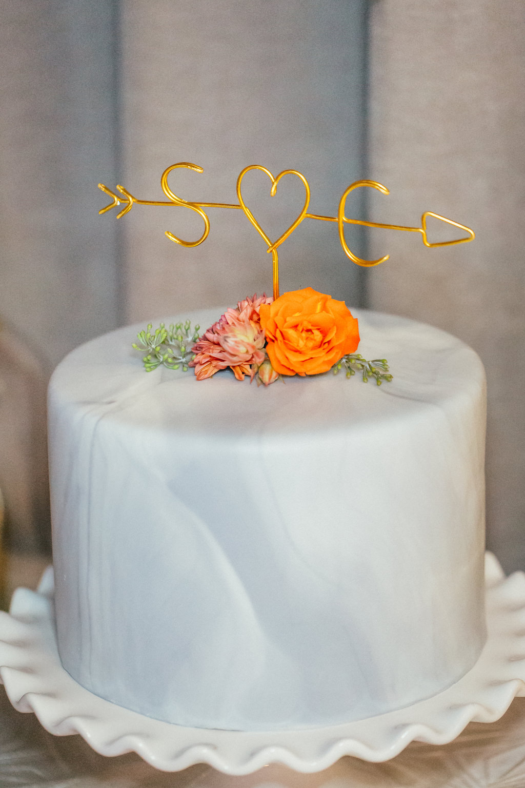 Single Tier Round Blue Marble Cake with Custom Initial and Arrow Heart Gold Wire Cake Topper and Orange Flowers on White Ceramic Cake Stand