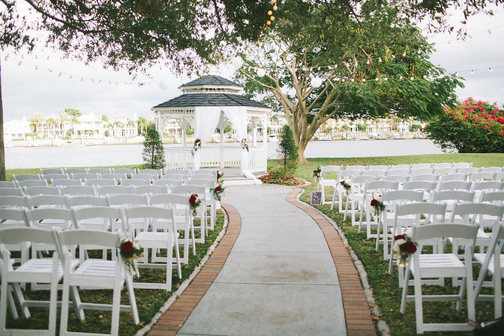 Outdoor Garden Wedding Ceremony Decor with White Wooden Folding Chairs, and Red and White Rose with Greenery Flowers with Twine, and Waterside Gazebo with White Draping | Tampa Wedding Ceremony Venue Davis Island Garden Club