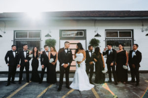Outdoor Industrial Modern Indian Wedding Party Portrait, Bridesmaids in Mismatched Black Floor Length Dresses, Groom in Black Tuxedo with White Boutonniere, Bride in Strapless Mermaid Wedding Dress with White Bouquet | Tampa Bay Wedding Photographer Grind and Press Photography