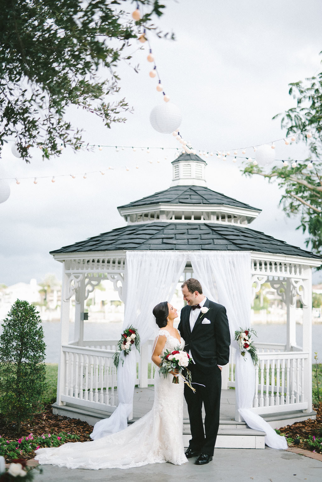 Outdoor Garden Wedding Ceremony Portrait, Bride and Groom in front of White Draped Gazebo with String LIghts and White Paper Lanters, White and Red Rose Florals and Bouquet with Greenery | Tampa Wedding Photographer Kera Photography | Waterfront Ceremony Venue Davis Island Garden Club