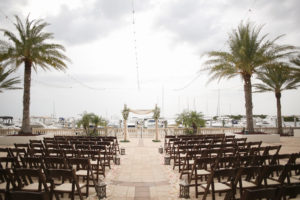 Rustic Nautical Outdoor Waterfront Marina Wedding Ceremony Decor with Brown Wood Folding Chairs, Rustic Hurricane Lanterns, Blush Pink Rose Petal Aisle, String Lights, and White Draped with Floral and Greenery Arch | Tampa Bay Wedding Venue The Westshore Yacht Club