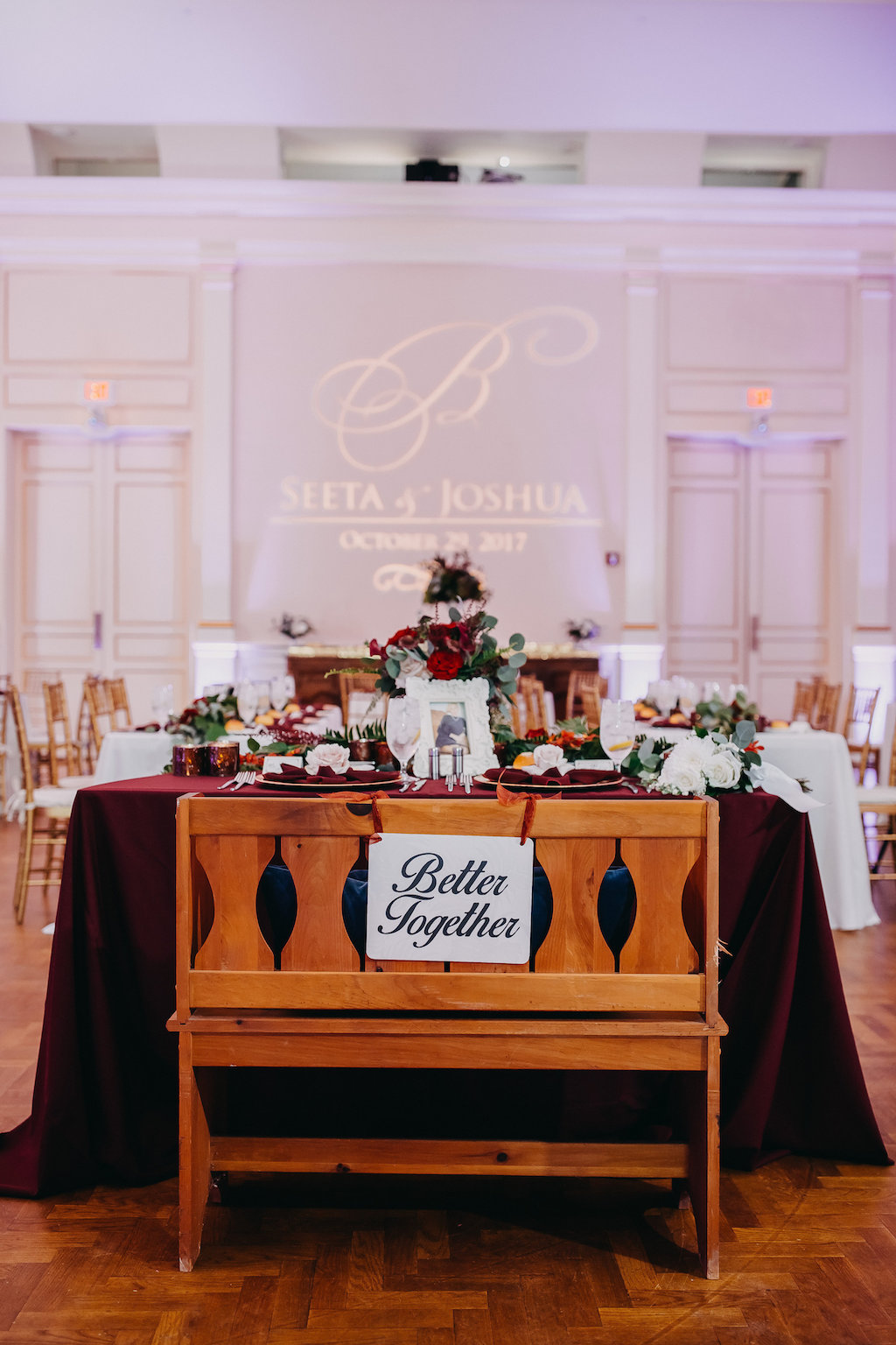 Ballroom Wedding Reception Sweetheart Table with Wooden Bench and Better Together Sign, Red Linens and Red and White with Greenery Centerpiece, and Custom Bride and Groom Name with Initial Gold Projection | Venue St Petersburg Museum of Fine Art