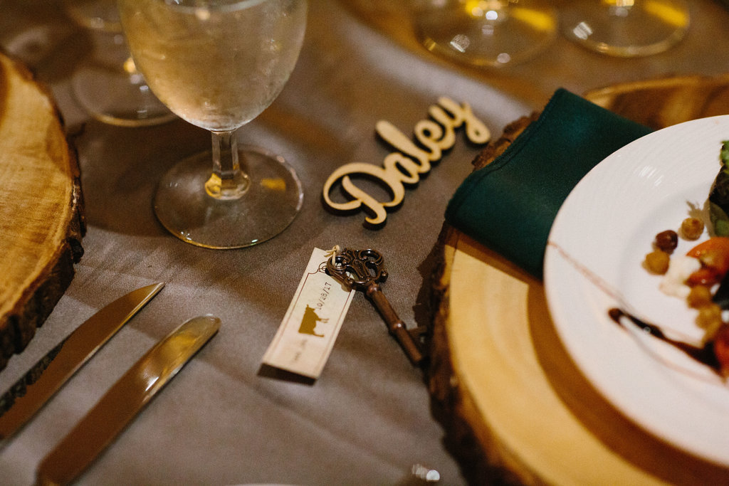 Natural Forrest Inspired Wedding Reception Table Decor with Natural Bark Edged Wooden Chargers, Forrest Green Napkins, Antique Key and Printed Paper Escort Card, and Laser Cut Wooden Name Card