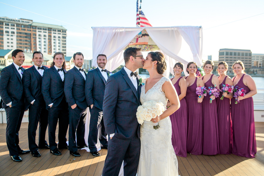 Outdoor On the Water Wedding Party Portrait, Bride in V Neck Lace Maggie Sottero Wedding Dress with White Rose Bouquet, Bridesmaids in V Neck Fuchsia Davids Bridal Dresses with Purple, Blue, and Red Boquets, Groom and Groomsmen in Navy Suits with Bow Ties | Tampa Unique Wedding Venue The Yacht Starship